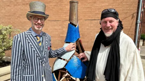 A man with glasses and a short grey beard is wearing a yellow- and black-striped top hat, with a pinstripe blazer over a waistcoat and a blue and yellow tie. On his left is a piece of art which looks like a cracked vase. On the other side of the artwork is a man with a black bandana and black scarf over a white tunic. Both men are resting their hands on the artwork.