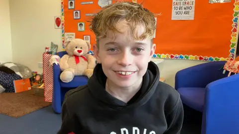 A young boy with blond wavy hair smiles at the camera. He is in a classroom with a bright red display board behind him.