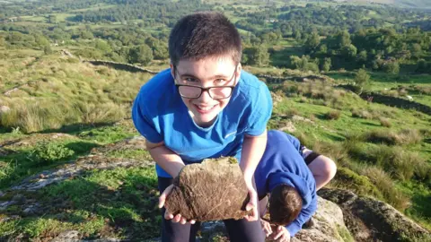 David Ridout 12-year-old Adam wearing a blue t-shirt with glasses and holding a rock on the top of a rocky hill.