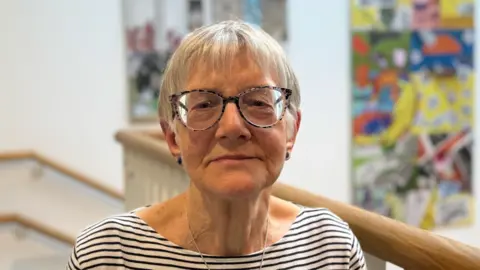 Elliot Deady/BBC A woman - Diana Minns - looks straight at the camera with a neutral expression. She is wearing a white and black striped top and glasses. There is abstract artwork on the wall behind her.