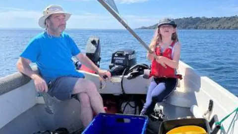 FAMILY PHOTO A small girl sits in a small boat with a middle aged man. The boat has an outboard motor and has an island behind. The girl holds a flag.