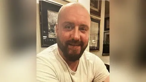 Pat Burns with short brown hair and beard wearing a white t-shirt sitting in a pub. He is smiling.