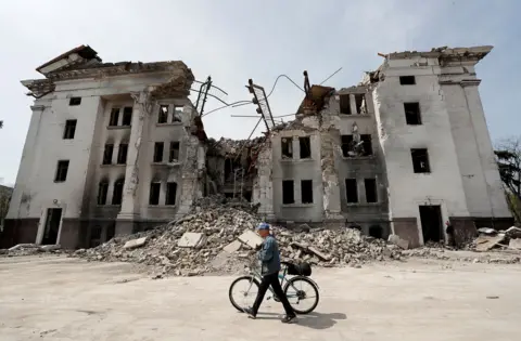 Alexander Ermochenko/Reuters man walks his bike on the street in front of the Mariupol Theater, damaged and half -relaxed surrounded by ruins against the clear blue sky, in Mariupol, East Ukraine, April 25, 2022.