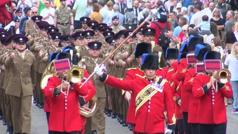 A large crowd surrounds columns of marching soldiers in brown uniforms in Newbury. At the front of the parade, a military band in red jackets and tall, black hats is led by a man holding up an ornate staff.