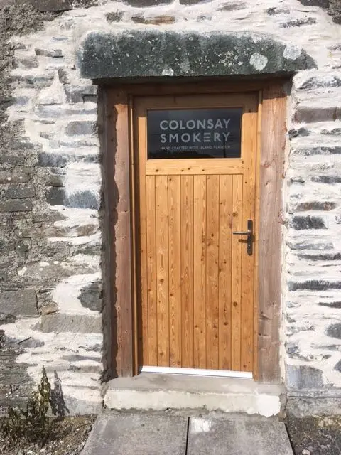 Richard Irvine A stone wall with a wooden door - it has a glass pane with the words "Colonsay Smokery" on it.