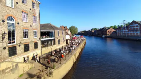 Buildings alongside a river. Crowds of people are sat on pub benches next to the river.