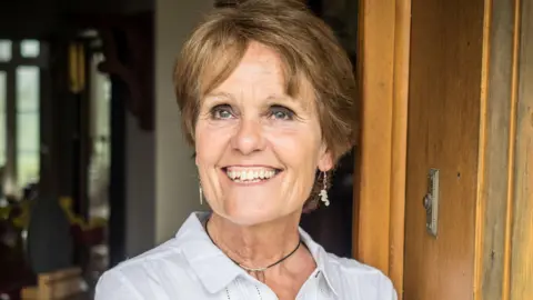 Supplied Gina Rippon. She has short brown hair and blue eyes. She is wearing a white linen shirt and black necklace, paired with seahorse shaped pendant earrings. She is leaning on a wooden door. She is smiling and looking at something out of shot.