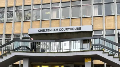 The exterior of Cheltenham Magistrates' Court. It is a beige building with light grey details and lots of windows. Two stairways can be seen either side of the entrance. Above the door there is a sign reading "Cheltenham Courthouse" in capital letters. 