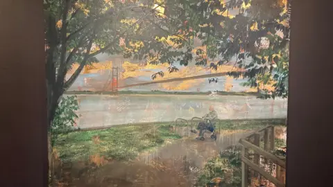 Emma Prempeh A photo of a painting hanging in a gallery, the paining portrays a man sat resting on a walking aid looking out towards the River Humber, with the Humber Bridge behind him. A tree frames the scene.