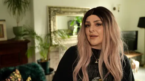 A woman with pink hair is looking to the side of camera. A living room decorated with plants and cushions is behind her out of focus.