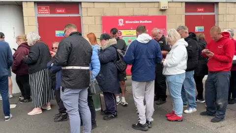 About two dozen people standing in a line outside the stadium. They are mainly looking at their phones. There are red doors behind them and a red sign which reads: "Riverside Stadium."