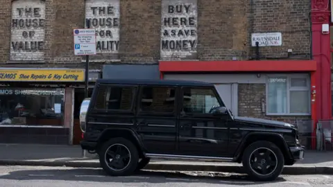 Getty Images A large black SUV is parked outside shops on Ravensdale Road in N36, including a shoe repair and key cutting business. There are Victorian style markings on the brickwork above the shops, advertising businesses that used to be there