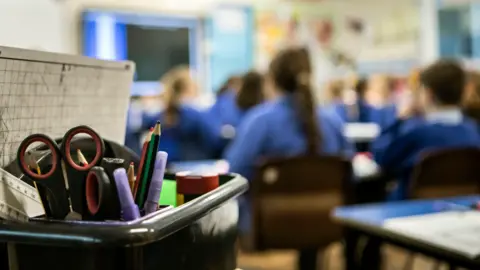 PA Media A classroom full of students who are out of focus - wearing blue shirts, with their backs to the camera. In the foreground is a box with stationery in it, including pencils and scissors.