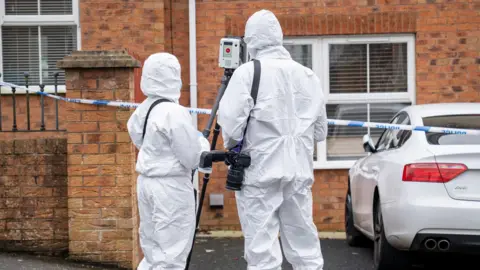 Aodhán Roberts Two forensic officers outside a red brick house. A camera is attached to a strap on one of their shoulders. A white car is parked outside. 