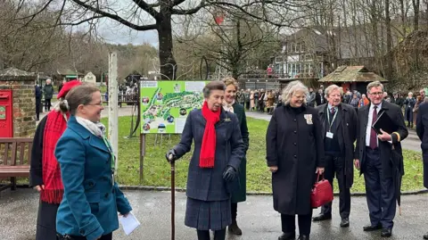 George Carden/BBC Princess Anne wearing a long, dark blue coat and red scarf. She is holding walking stick in her right hand and is surrounded by half a dozen people. They are on a driveway with green grass and a tree behind them. there is a red post box built into a brick wall to the left. The sky is grey