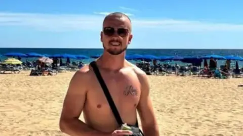 Family picture Nathan wearing sunglasses and standing on a sandy beach, with the sea behind him. He is not wearing a shirt, with a black bag strap across his chest. In the background there is a row of blue beach umbrellas and sun loungers. 