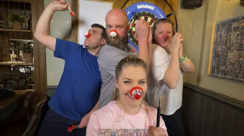 A man in a blue T-shirt is side on to the camera on the left. Another man and two women are facing the camera and all four have red noses on.