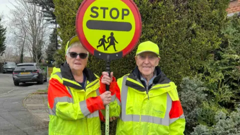 Reg Bown Reg and Anne, both wearing high-vis, with the stop sign between them, are pictured standing at the crossing