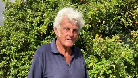 A elderly man wearing a dark blue polo shirt and looking at the camera. He is standing in front of a green bush