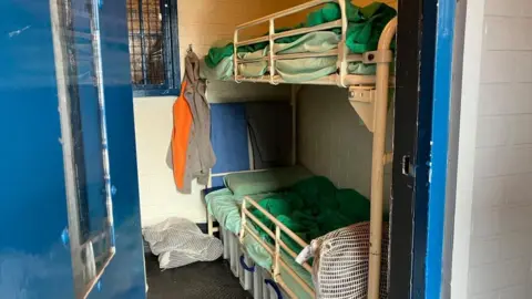 BBC/ Nicola Rees The door of a prison cell is ajar. Inside a bunk bed can be seen made up with green blankets and pillows and with guard rails on the sides .