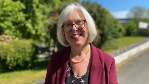 Dr Nicola Brink stands outdoors on a sunny day. Her hair is in a grey bob, and she is wearing a burgundy jacket and glasses. She stands on a path with greenery and a low wall in the background.