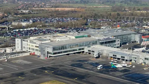 Aerial view of Newcastle Airport. There is a large terminal building with giant windows and a plane parked outside. In the distance are car parks full of vehicles.