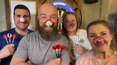 Two men on the left and two women on the right are looking at the camera. Three of them are holding darts on the photo and a dartboard is partly visible in the background.