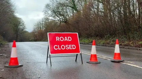 A red road closed sign which has one traffic cone to the left of it and two on the right. The sign is placed in the middle of the road. There are trees lining both sides of the road. 