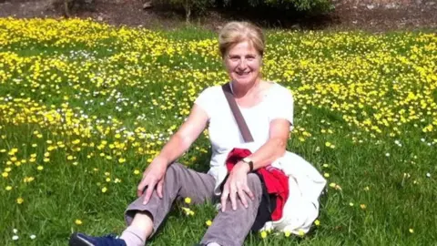 Wiltshire Police Danielle Carr-Gomm sitting in a grassy field surrounded by buttercups, smiling at the camera. She is wearing grey trousers and a white t-shirt, with a cross body bag.