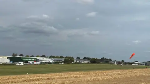 A view of the airfield under a grey sky. A mown yellow crop field with a green airstrip next to it. Aviation buildings in the background. One small plan on the grass airstrip.