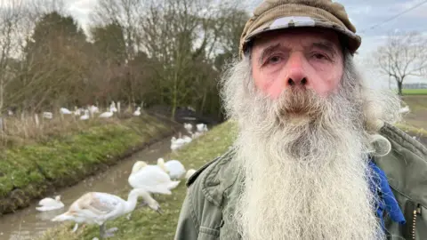 David Elston has a long grey beard and brown moustach. He's wearing a flat cap and is surrounded by swans. 