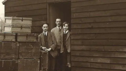 Wiltshire & Swindon History Centre An old black & white picture showing the doorway of a large wooden hut with three men stood in it, dressed smartly in jackets and ties. There are a stack of trunks and boxes outside, presumably filled with books