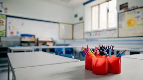 Getty Images An empty school classroom with tubs of pencils on a desk and an array of seats and tables