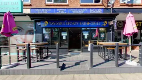 The front of a premises called 'Taylor's Sports Bar and Grill', as indicated by a blue sign above the glass door. Tables and parasols sit on a small decked area outside.