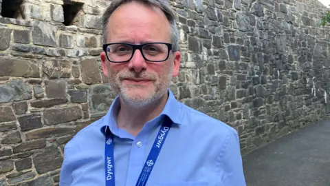 Philip Kloer, in a blue shirt and blue lanyard, with glasses and grey hair and stubble in front of a stone wall