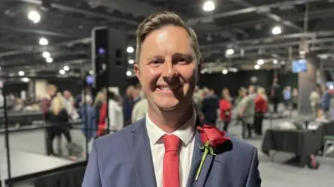 A white man in his 30s with light brown hearing.  He is wearing a mid-blue blazer with a white shirt and red tie.  He has a large red rose on his lapel.
