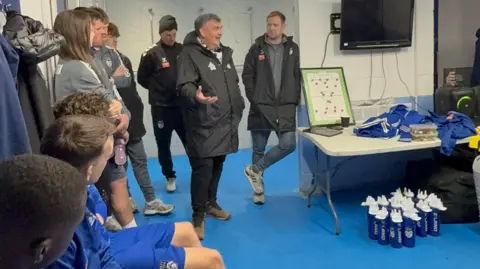 Prostate Cancer UK The image shows the inside of a football changing room. Several individuals are present, some seated on benches along one wall and others standing near the entrance to an adjoining shower or corridor area.