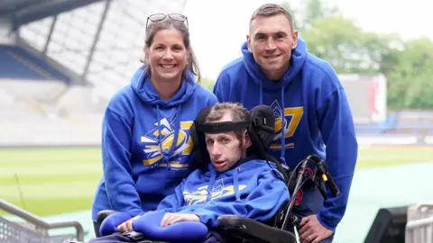 Kevin Sinfield, Rob Burrow and Lindsey Burrow look at the camera at a sport stadium. All three are wearing blue hooded tops. 