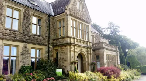 Google The exterior entrance of Ullenwood Manor. It is a large sand coloured building with tall windows and an arched doorway, with various bushes and plants outside.