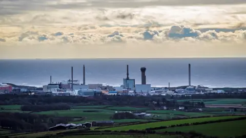 A general view of Sellafield taken from afar across a green field. The skyline of Sellafield is characterized by tall buildings and towers, with the sea in the background. 