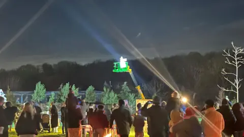 John Devine/BBC A figure in white is standing on top of a telehandler's arm and platform, about six metres (20ft) in the air. A crowd of people are in the foreground, some with camera phones. Spotlights illuminate the figure and conifer trees can be seen behind it.