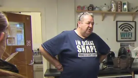Gerard Woodhouse, wearing a blue t-shirt that says 'In great shape on it' in white capital letters, is standing in the kitchen of a community centre 