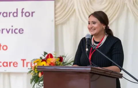 Nishkam Healthcare Trust A woman in black pinstripe dress with a pearl necklace and long dark hair tied back smiles as she stands in front of a lectern with microphone. A marquee side can be seen behind her and part of an award banner. A flower stand has yellow red and orange blooms.