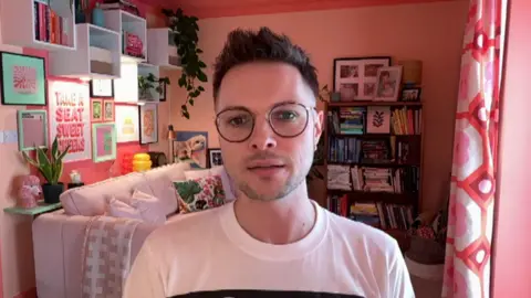 Nathaniel has short, dark hair spiked up at the front. They wear round glasses and a white t-shirt. Their room is painted pink and decorated with pictures organised in a gallery wall behind their pink couch