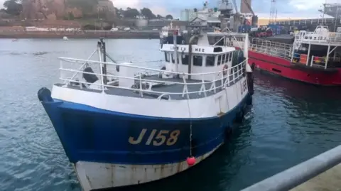 A white and blue fishing boat with J158 on the left side, in the ocean, next to another vessel in the harbour. Roads and a hill can be seen in the background.