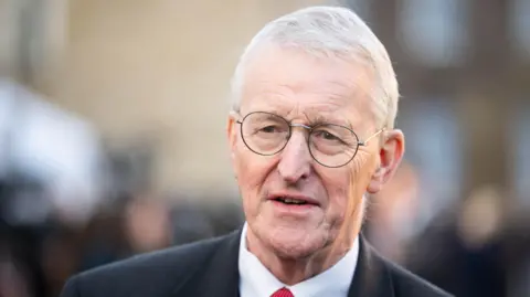 A headshot of an older man with grey hair wearing glasses. He is standing outside. The background is blurred.