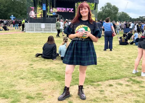 Blaize Ashton-Jenkins Blaize is stood in front of a stage at a festival wearing a Guns 'N' Roses band t-shirt. She is smiling. 