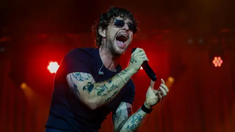 Tom Grennan singing on stage. He holds a microphone and his mouth is wide open. He wears dark glasses and a black T-shirt and has tattoos on his arms. 