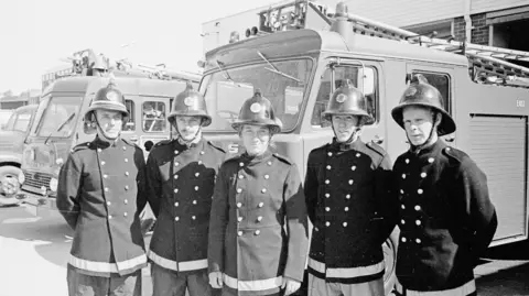 East Sussex Fire and Rescue Service A young Mary Joy Langdon in the centre in a firefighters uniform, flanked by two male colleagues in uniform on either side. They are stood in front of a row of 1970s fire engines in the black and white image.