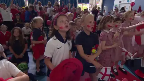 Many children are standing up looking at a TV.